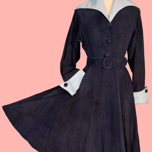 May include: A vintage black coat with a light blue collar and cuffs. The coat features a belted waist, long sleeves, and a flared skirt. The coat is displayed on a mannequin against a pink background.