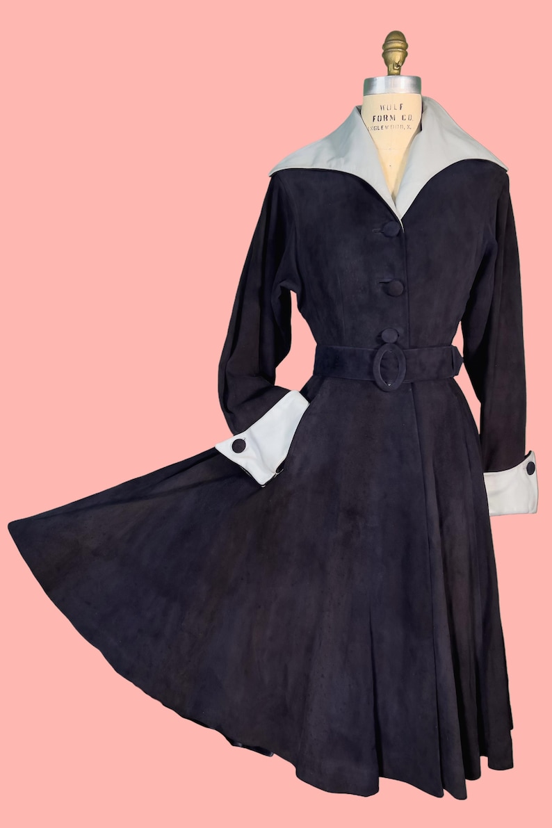 May include: A vintage black coat with a light blue collar and cuffs. The coat features a belted waist, long sleeves, and a flared skirt. The coat is displayed on a mannequin against a pink background.