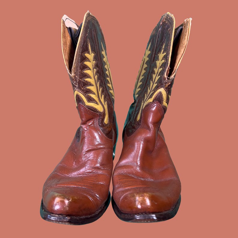 May include: A pair of brown leather cowboy boots. The boots have a pointed toe and a decorative design in yellow and brown on the upper shaft. The boots are set against a solid orange background.