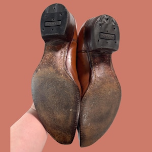 May include: A pair of brown leather cowboy boots, viewed from the soles up. The soles are worn, showing signs of use. The heels are dark brown with the brand name "Solerich" embossed on the rubber. The leather uppers are a lighter brown.