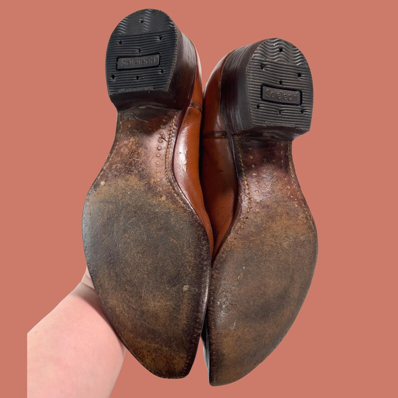 May include: A pair of brown leather cowboy boots, viewed from the soles up. The soles are worn, showing signs of use. The heels are dark brown with the brand name "Solerich" embossed on the rubber. The leather uppers are a lighter brown.