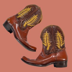 May include: A pair of brown leather cowboy boots with yellow decorative stitching on the upper shaft. The boots have a pointed toe and a stacked heel, showcasing a classic Western style.