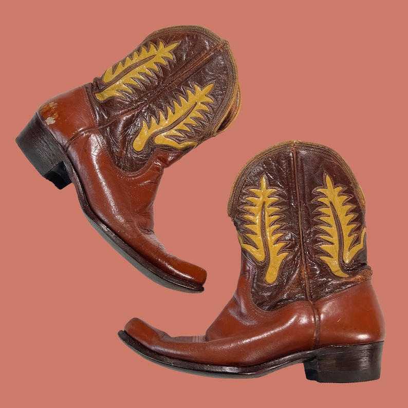 May include: A pair of brown leather cowboy boots with yellow decorative stitching on the upper shaft. The boots have a pointed toe and a stacked heel, showcasing a classic Western style.