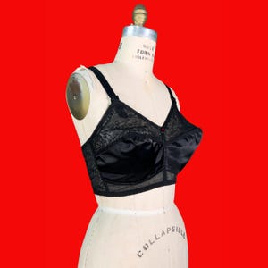 May include: Black vintage bra with lace detailing and satin cups. The bra is displayed on a mannequin against a red background. The bra has adjustable straps and a front closure.