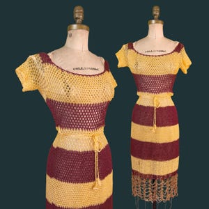 May include: Two vintage crochet dresses on mannequins. The dresses have horizontal stripes in yellow and burgundy. The short-sleeved dresses have a scoop neckline and a matching belt. One dress has a fringed hemline.