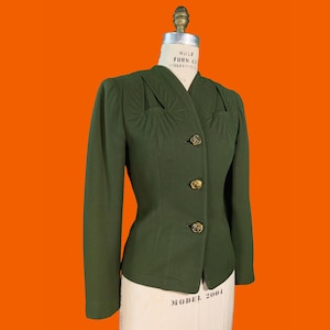 May include: An olive green long-sleeve jacket featuring a V-neckline, three gold-toned circular buttons down the front, and decorative radiating pleats at the upper chest area. The jacket is displayed on a white dress form with visible text.