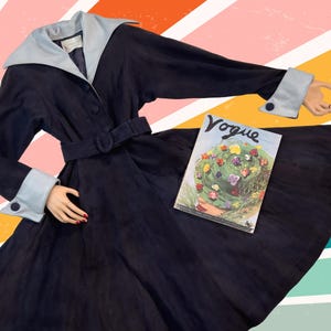 May include: A vintage navy blue coat with a light blue collar and cuffs. The coat has a matching belt and button details. A vintage Vogue magazine is placed on the coat. The background has a colorful striped design.