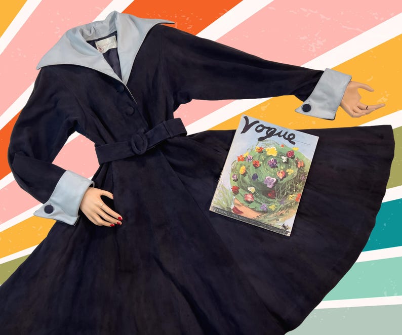 May include: A vintage navy blue coat with a light blue collar and cuffs. The coat has a matching belt and button details. A vintage Vogue magazine is placed on the coat. The background has a colorful striped design.
