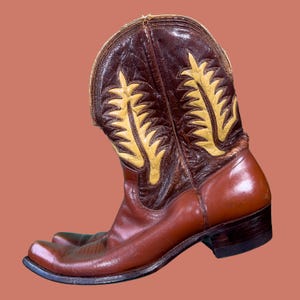 May include: A brown and tan cowboy boot with a pointed toe. The boot features decorative yellow stitching and a leather upper. The boot is approximately 12 inches tall.