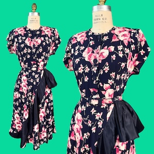 Può includere: Un abito blu navy con motivo floreale rosa e bianco, maniche corte, scollo rotondo e chiusura frontale con bottoni. L'abito include una cintura abbinata in vita e un dettaglio a balza in tessuto blu navy sul davanti della gonna.