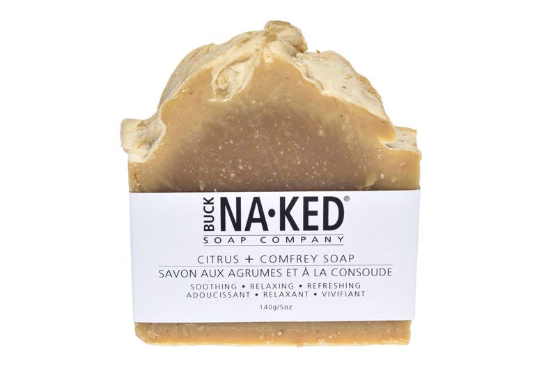 Może przedstawiać: Kostka mydła w jasnobrązowym kolorze o teksturowanej powierzchni. Mydło ma białą etykietę z napisem "BUCK NA-KED SOAP COMPANY" i "CITRUS + COMFREY SOAP". Etykieta zawiera r&oacute;wnież tekst "SOOTHING &bull; RELAXING &bull; REFRESHING" i "140g/5oz".