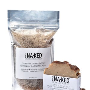 May include: A clear plastic bag filled with brown coffee-scented Dead Sea salt soak. The bag has a white label with the text "BUCK NA-KED SOAP COMPANY" and "COFFEE START UP DEAD SEA SOAK INFUSION AUX SELS DE LA MER MORTE" in black. The bag is sitting on a white surface with coffee beans scattered around it. A brown bar of coffee-scented soap with the text "BUCK NA-KED SOAP COMPANY" and "COFFEE START UP SOAP SAVON &Eacute;NERGIE MATINALE AU CAF&Eacute;" in black is sitting on the right side of the image.
