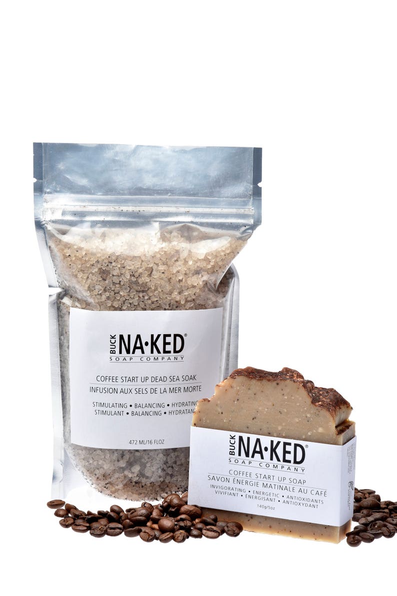 May include: A clear plastic bag filled with brown coffee-scented Dead Sea salt soak. The bag has a white label with the text "BUCK NA-KED SOAP COMPANY" and "COFFEE START UP DEAD SEA SOAK INFUSION AUX SELS DE LA MER MORTE" in black. The bag is sitting on a white surface with coffee beans scattered around it. A brown bar of coffee-scented soap with the text "BUCK NA-KED SOAP COMPANY" and "COFFEE START UP SOAP SAVON &Eacute;NERGIE MATINALE AU CAF&Eacute;" in black is sitting on the right side of the image.