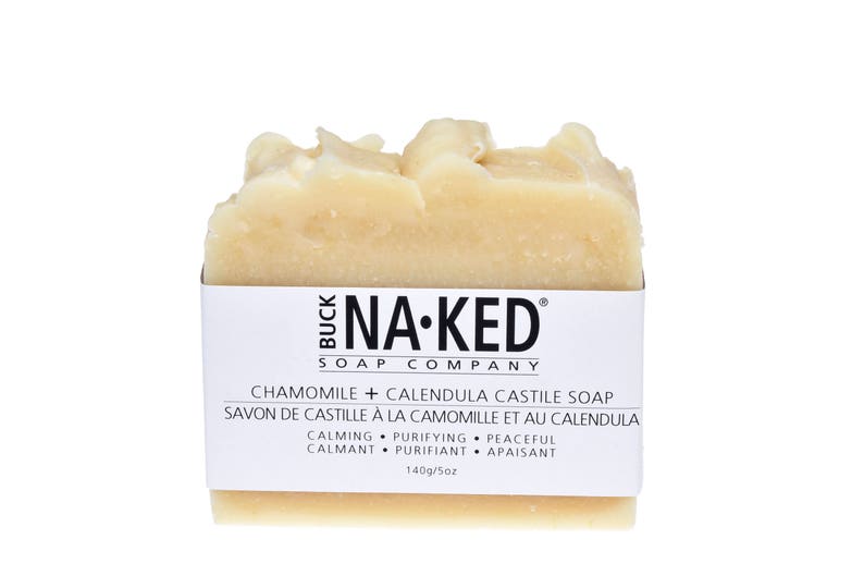 May include: A bar of white soap with the text "NA-KED SOAP COMPANY" and "CHAMOMILE + CALENDULA CASTILE SOAP" printed on the label. The soap is labeled as calming, purifying, and peaceful.