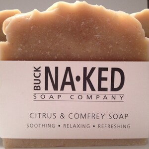 Może przedstawiać: Kostka mydła cytrusowego i żywokostu z Buck Naked Soap Company. Mydło jest jasnobrązowe z teksturowaną g&oacute;rą. Etykieta głosi "Citrus & Comfrey Soap" ze słowami "Soothing, Relaxing, Refreshing" poniżej.