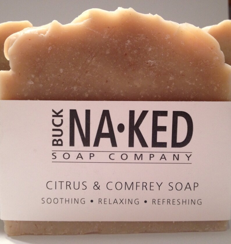 Może przedstawiać: Kostka mydła cytrusowego i żywokostu z Buck Naked Soap Company. Mydło jest jasnobrązowe z teksturowaną g&oacute;rą. Etykieta głosi "Citrus & Comfrey Soap" ze słowami "Soothing, Relaxing, Refreshing" poniżej.