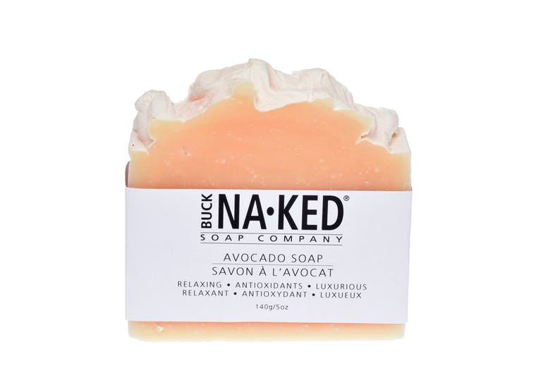 Puede incluir: Una pastilla de jab&oacute;n de aguacate de Buck Na-Ked Soap Company. El jab&oacute;n es de color melocot&oacute;n claro y tiene una etiqueta blanca con texto negro que dice "Avocado Soap, Savon &agrave; l'avocat, Relaxing, Antioxidants, Luxurious, Relaxant, Antioxydant, Luxe, 140g/5oz."