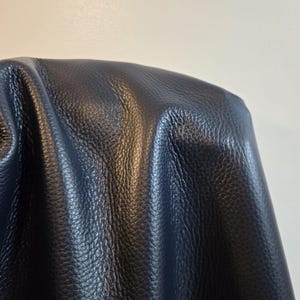 Può includere: Un pezzo di pelle blu navy scuro testurizzato con un motivo a grana visibile. Il materiale è drappeggiato, creando pieghe morbide e riflessi che enfatizzano la consistenza granulosa della superficie e il colore profondo e saturo del materiale.
