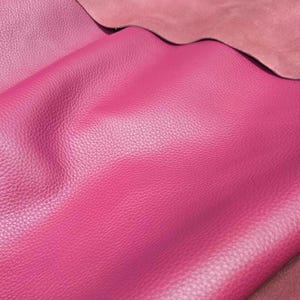 Pode incluir: Close-up de duas peças de couro em tons de rosa. A peça superior tem uma textura mais clara, semelhante a camurça, enquanto a inferior é um magenta vibrante e texturizado. O couro parece macio e flexível, adequado para artesanato.