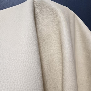 Può includere: Un pezzo di pelle color bianco sporco caratterizzato da una distinta texture granulata. Il materiale è drappeggiato per mostrare la sua flessibilità e superficie morbida, evidenziando il motivo naturale della grana e la finitura opaca della pelle.