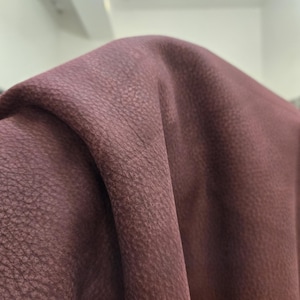 Può includere: Primo piano di un tessuto drappeggiato di colore bordeaux scuro. Il materiale ha un aspetto strutturato, simile alla pelle, con sottili variazioni di tono, creando un'estetica ricca e tattile. Le pieghe e le ombre aggiungono profondità all'immagine.
