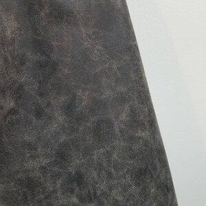 Op de afbeelding: Close-up van een donkergrijze, textuurrijke stof met een licht verweerde uitstraling. De stof heeft een subtiele glans en een licht ruw gevoel.