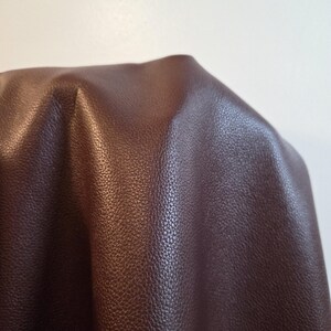 Può includere: Un pezzo di tessuto in similpelle marrone scuro con una distinta consistenza granulosa. Il materiale è drappeggiato per mostrare la sua flessibilità e la lucentezza morbida, evidenziando il motivo a grana coerente sulla superficie del materiale sintetico.