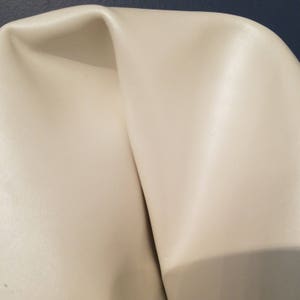 Pode incluir: Close-up de um tecido de couro liso de cor branco-queimado. O tecido tem um brilho sutil e uma textura macia e flexível.