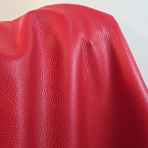 Può includere: Un materiale in pelle rosso vibrante con una distinta consistenza granulata. La superficie mostra pieghe e grinze naturali, creando ombre e riflessi morbidi sul materiale. La pelle ha una finitura opaca e un colore rosso profondo e uniforme.