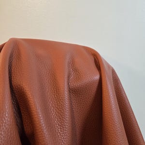 Può includere: Primo piano di un foglio di pelle marrone con superficie strutturata. La pelle è drappeggiata, mostrando pieghe e ombre. Il colore è un marrone rossastro caldo.