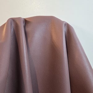 Può includere: Un pezzo di pelle color malva con una distinta trama granulata. Il materiale è drappeggiato per creare morbide pieghe e ombre, evidenziando la finitura opaca e il motivo a grana coerente su tutta la superficie del materiale flessibile.