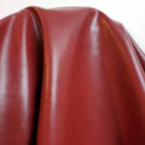 Pode incluir: Close-up de um tecido vermelho escuro, liso e semelhante ao couro. O tecido está drapeado sobre uma superfície, criando dobras e curvas suaves.