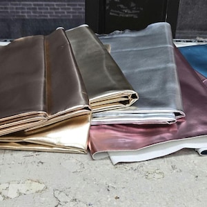 Può includere: Campioni di tessuto metallizzato piegati in vari colori: bronzo, oro, argento, rosa e verde acqua. I tessuti sono impilati su una superficie chiara, mostrando la loro texture lucida.