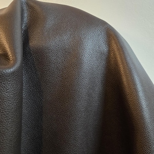 Può includere: Un materiale marrone scuro con una distinta texture granulata. La superficie presenta una finitura opaca con sottili riflessi dove la luce colpisce le pieghe e le increspature del tessuto, creando un aspetto morbido e drappeggiato nell'inquadratura.