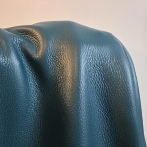 Può includere: Un pezzo di pelle color verde acqua scuro con una texture a grana naturale visibile. Il materiale è drappeggiato per creare pieghe e riflessi morbidi, mettendo in risalto la finitura opaca e il colore blu-verde profondo e saturo della superficie.