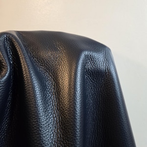 Può includere: Un materiale blu scuro con una distinta consistenza granulata. La superficie presenta pieghe e increspature profonde che creano ombre e riflessi naturali, mettendo in risalto la qualità flessibile e morbida del materiale blu scuro.