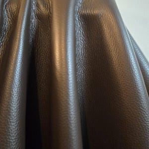 Può includere: Un pezzo di pelle marrone scuro con una distinta trama granulata. Il materiale è drappeggiato in pieghe verticali, creando ombre e riflessi profondi sulla superficie che enfatizzano la grana e la natura flessibile della pelle.