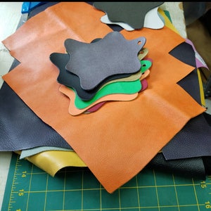 May include: A stack of different colored leather pieces, including orange, black, green, and gray. The leather pieces are arranged in a star shape and are on a green cutting mat.