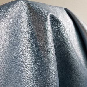 May include: Close-up of a textured, blue leather fabric with a slight sheen. The fabric is folded and draped, showcasing its texture and color.