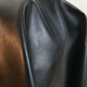 May include: A close-up of black faux leather fabric with a slight sheen. The fabric is folded and draped, showing its texture and drape.