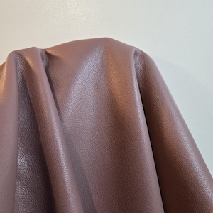 Può includere: Un pezzo di pelle color malva con una distinta consistenza granulata. Il materiale è drappeggiato per mostrare la sua flessibilità e la superficie morbida, evidenziando il motivo a grana coerente e il tono viola polveroso e tenue della pelle.