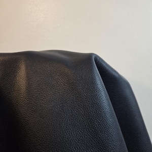 Può includere: Un pezzo di pelle blu scuro con una distinta consistenza granulata. Il materiale è ripiegato, creando curve morbide e ombre che mettono in risalto la grana e la finitura opaca della superficie sulla sezione visibile della pelle.