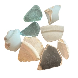 Puede incluir: Una colección de fragmentos de vidrio marino y cerámica de diversas formas y tamaños. Las piezas son de tonos verdes, blancos y beige, algunas con patrones y texturas. Los fragmentos están dispersos sobre un fondo blanco.
