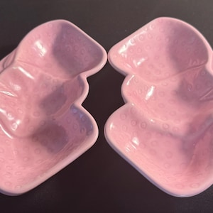 Puede incluir: Dos platos rosas de cerámica con forma de fresa. Cada plato tiene una superficie texturizada con pequeños círculos espaciados uniformemente. Los platos están apilados y colocados sobre una superficie oscura. Estos platos decorativos son ideales para servir aperitivos.