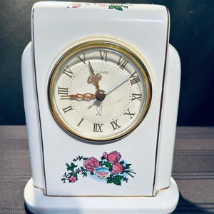 May include: White porcelain clock with a gold rim and Roman numeral face. The clock has a floral design with pink roses and green leaves. The clock face reads "QUARTZ" and "S" for seconds.