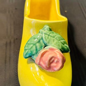 May include: A yellow ceramic planter shaped like a shoe with a pink rose and green leaves on the side.