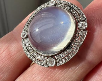 Op de afbeelding: Een ovale antieke broche met een grote, doorschijnende, lichtblauwe edelsteen in het midden. De edelsteen is gezet in een gedetailleerde zilveren setting, bezet met kleine, sprankelende diamanten. De broche heeft een klassiek, vintage ontwerp.