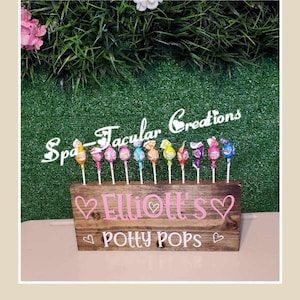 May include: A wooden sign with the text "Elliot's Potty Pops" in pink lettering with hearts. The sign has a row of colorful lollipops attached to it. The sign is on a green surface.