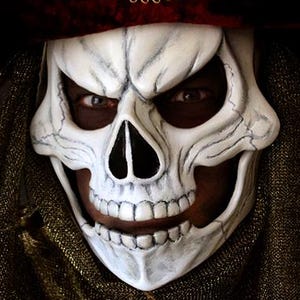 Peut inclure: Un costume de théâtre avec un masque de crâne blanc à l'expression menaçante. Le masque est associé à un chapeau noir et rouge foncé orné de plumes et de garnitures dorées. Le costume comprend un col or et bordeaux.