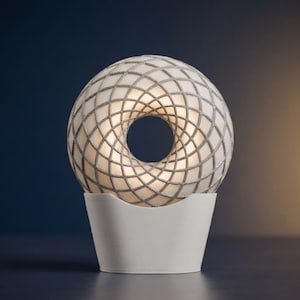 Può includere: Una lampada sferica con un centro cavo e un esterno bianco con finitura opaca, caratterizzata da un motivo a griglia incrociata in rilievo color argento metallizzato. La lampada è sostenuta da una base solida a forma di coppa, bianca e opaca.